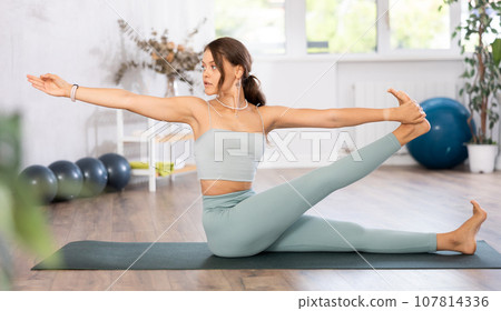 Young woman yoga trainer performing Revolved Seated Hand to Big Toe pose on black rug Young woman yoga trainer performing Revolved Seated Hand to Big Toe pose on black rug 107814336