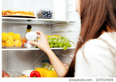 Woman hand taking, grabbing or picks up camembert or brie cheese out of open refrigerator shelf, fridge drawer full of fruits, vegetables, banana, peaches, yogurt. Healthy food diet, lifestyle concept Woman hand taking, grabbing or picks up camembert or brie cheese out of open refrigerator shelf, fridge drawer full of fruits, vegetables, banana, peaches, yogurt. Healthy food diet, lifestyle concept 107814609