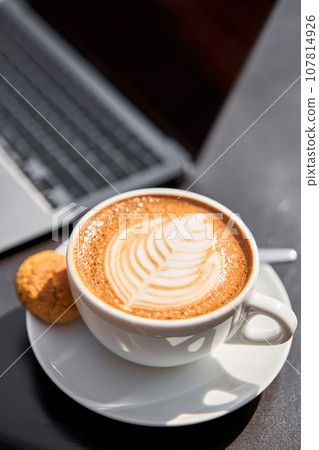 A cup Capuchino coffee. The table in the coworking space of the coffee shop. A cup of coffee on the veranda on a sunny day. Coffee break at the cafe. Close-up A cup Capuchino coffee. The table in the coworking space of the coffee shop. A cup of coffee on the veranda on a sunny day. Coffee break at the cafe. Close-up 107814926