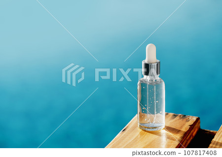 One mockup transparent glass bottle on wooden deck over blue water. cosmetics collagen serum, organic essential or aroma oil for beauty skin care. Summer hydrating and vitalizing essentials 107817408