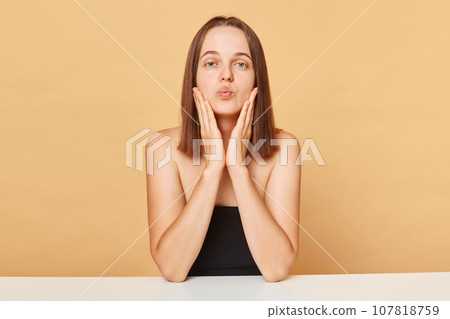 Romantic attractive Caucasian female model wearing black top sitting at table isolated over beige background posing with pout lips sending air kissing. Romantic attractive Caucasian female model wearing black top sitting at table isolated over beige background posing with pout lips sending air kissing. 107818759