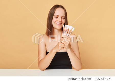 Beautiful brown haired woman with make-up brushes near attractive face smiling while looking away wearing black top posing with bare shoulders isolated over beige background. Beautiful brown haired woman with make-up brushes near attractive face smiling while looking away wearing black top posing with bare shoulders isolated over beige background. 107818760