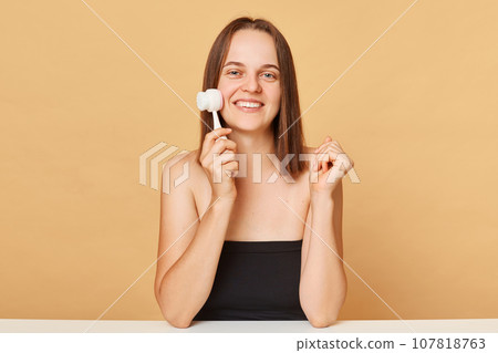 Young smiling woman using face cleansing brush enjoying facial beauty procedures and skin care morning routine wearing black top isolated over beige background. Young smiling woman using face cleansing brush enjoying facial beauty procedures and skin care morning routine wearing black top isolated over beige background. 107818763