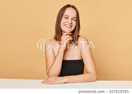Confident smiling Caucasian female model wearing black top sitting at table isolated over beige background holding her chin looking with satisfied expression. Confident smiling Caucasian female model wearing black top sitting at table isolated over beige background holding her chin looking with satisfied expression. 107818765