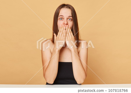 Shocked surprised Caucasian female model wearing black top sitting at table isolated over beige background looking at something astonishing covering mouth with palms. Shocked surprised Caucasian female model wearing black top sitting at table isolated over beige background looking at something astonishing covering mouth with palms. 107818766