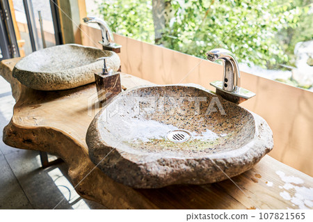 Stone beautiful sink with a metal faucet next to an mirror. Close-up of an elegant faucet in the bathroom sink next to large windows. Stone beautiful sink with a metal faucet next to an mirror. Close-up of an elegant faucet in the bathroom sink next to large windows. 107821565