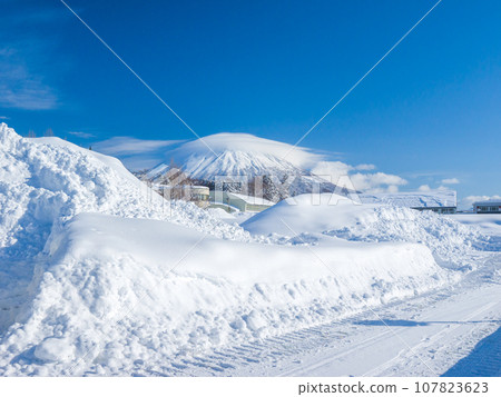 View of the volcano covered in clouds beyond the snow pile on the town corner (Niseko Town, Hokkaido) 107823623