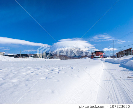 View of houses and the volcano covered in clouds beyond the snowfield (Niseko Town, Hokkaido) 107823630