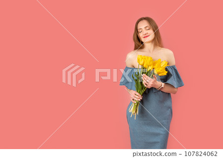 Beautiful young woman with closed eyes in bare-shouldered dress holding bouquet of yellow flowers, pink background. Copy space, 107824062