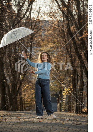 Young woman with transparent umbrella happy smiles. Student with curly hair poses in autumn park 107824401