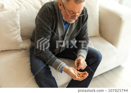 Middle aged senior man holding medical pills sitting on couch at home. Mature old senior grandfather taking medication cure pill vitamin. Age prescription medicine healthcare therapy concept 107825744