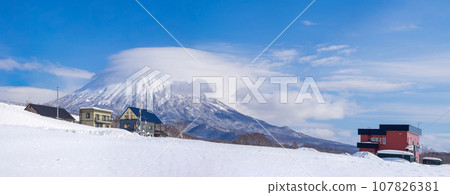 View of houses and the volcano covered in clouds beyond the snowfield (Niseko Town, Hokkaido) 107826381