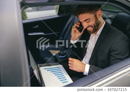 Smiling male analyst working on laptop and talking on phone while going to airport by car 107827215