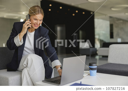 Smiling business woman working on laptop while talking phone sitting on background of modern office 107827216