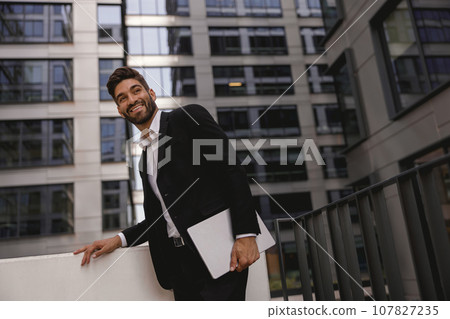 Stylish male manager standing with laptop on office terrace and looks away. High quality photo 107827235