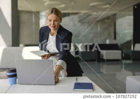 Smiling business lady in suit working on laptop sitting on background of modern office hall Smiling business lady in suit working on laptop sitting on background of modern office hall 107827252
