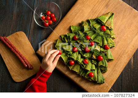 A little boy decorates a Christmas tree made of pancakes with cherry tomatoes and sausage. Pancake board A little boy decorates a Christmas tree made of pancakes with cherry tomatoes and sausage. Pancake board 107827324
