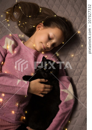 A teenage girl lies on a bed with a black cat wrapped in a garland, top view A teenage girl lies on a bed with a black cat wrapped in a garland, top view 107827332