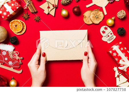Top view of woman holding an envelope on red background made of holiday decorations. Christmas time concept Top view of woman holding an envelope on red background made of holiday decorations. Christmas time concept 107827738
