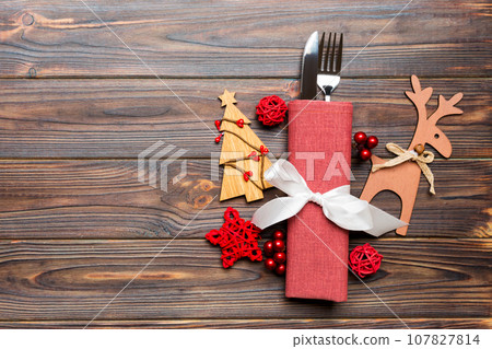 Top view of holiday objects on wooden background. Utensils tied up with ribbon on napkin. Christmas decorations and reindeer with copy space. New year dinner concept Top view of holiday objects on wooden background. Utensils tied up with ribbon on napkin. Christmas decorations and reindeer with copy space. New year dinner concept 107827814
