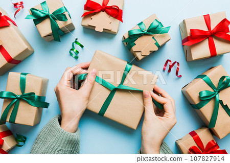 Woman is packing surprise gift for Christmas holidays, sitting at workplace with craft tools, top view. Woman wrapping Christmas gift at colored table Woman is packing surprise gift for Christmas holidays, sitting at workplace with craft tools, top view. Woman wrapping Christmas gift at colored table 107827914