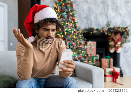 Man sitting alone on sofa for Christmas at home, hispanic received online notification on phone message with bad news, sad man wearing red santa hat in living room. 107828231