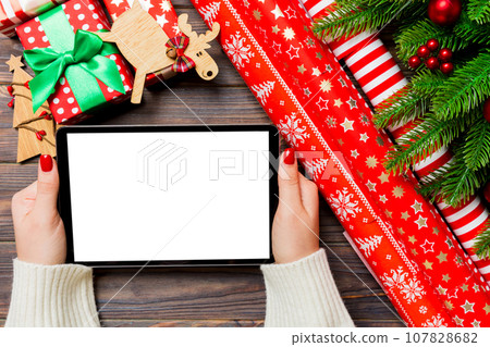Top view of female hands holding a tablet on wooden Christmas background made of fir tree and festive decorations. New year holiday concept. Mockup Top view of female hands holding a tablet on wooden Christmas background made of fir tree and festive decorations. New year holiday concept. Mockup 107828682