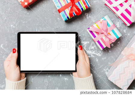 Top view of female hands holding a tablet on cement Christmas background made of gift boxes. New year holiday concept. Mockup 107828684