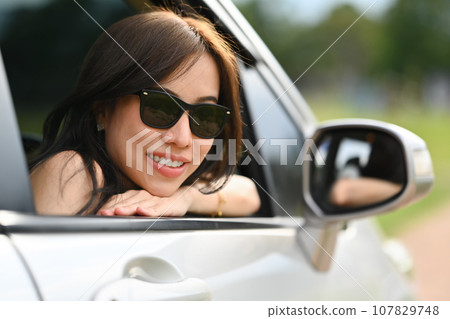 Attractive millennial woman in sunglasses leaning out of the car window enjoying the sunset. Active lifestyle, road trip and journey concept 107829748