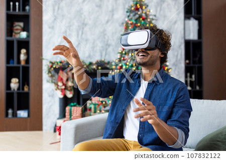 A man is using a virtual reality simulator for Christmas, sitting on a sofa in the living room of the house, near a decorated Christmas tree, on New Year's holidays. 107832122