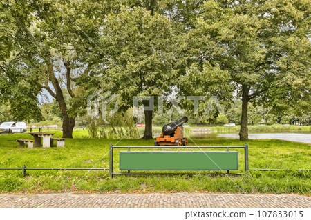 a green sign in the middle of a park with trees and grass on either side, there is a white van parked next to 107833015