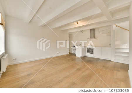 an empty living room with wood flooring and white painted walls in the corner to the kitchen is on the right 107833086