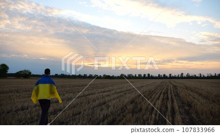 Ukrainian man walking on barley meadow with a blue-yellow banner on shoulders at sunset. Young guy going on wheat field with a flag of Ukraine as symbol of victory against russian aggresion. Slow mo 107833966