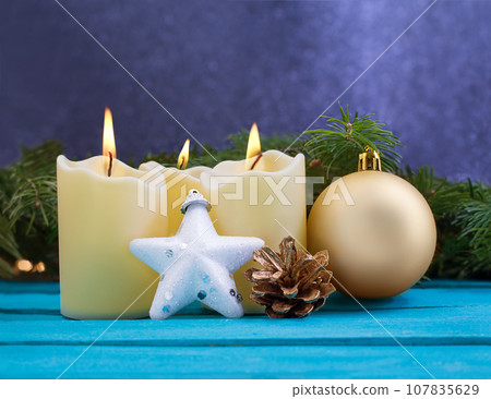 Christmas candles with fire, white sparkling star, golden pine cone and ball on blue wooden background. Behind there are spruce branches. New Year. Copy space Christmas candles with fire, white sparkling star, golden pine cone and ball on blue wooden background. Behind there are spruce branches. New Year. Copy space 107835629