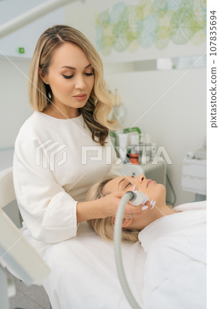 Vertical portrait of cosmetologist doing facial skin therapy. Radiofrequency face lifting. Hardware antiaging procedure. RF lifting and vacuum massage. Concept of non-surgery cosmetology. 107835694