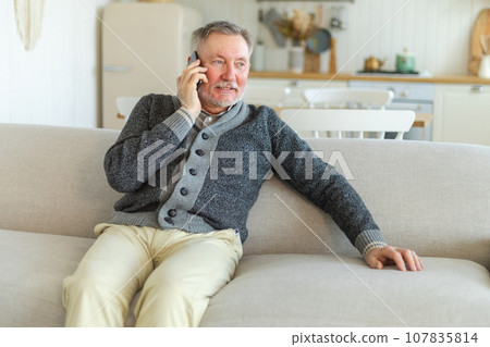 Happy middle aged senior man talking on smartphone with family friends. Older mature grandfather with cell phone talking with grown up children, resting at home. Older generation modern tech usage Happy middle aged senior man talking on smartphone with family friends. Older mature grandfather with cell phone talking with grown up children, resting at home. Older generation modern tech usage 107835814