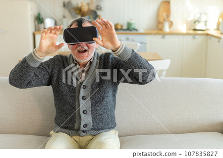 Excited mature senior man wearing using virtual reality metaverse VR glasses headset at home. Grandfather touching air during VR experience on virtual reality helmet. Simulation hi-tech videogame Excited mature senior man wearing using virtual reality metaverse VR glasses headset at home. Grandfather touching air during VR experience on virtual reality helmet. Simulation hi-tech videogame 107835827