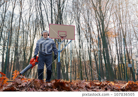 Handsome senior man removing dry leaves with leaf blower on basketball court outside. Low angle view of smiling bearded male worker with portable leaf blower working in morning. Seasonal work concept. Handsome senior man removing dry leaves with leaf blower on basketball court outside. Low angle view of smiling bearded male worker with portable leaf blower working in morning. Seasonal work concept. 107836505
