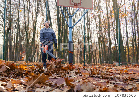 Qualified maintenance worker taking care of city park area during autumn season. Low angle view of aged man in overalls removing dry leaves with leaf blower at basketball court. Seasonal work concept. 107836509