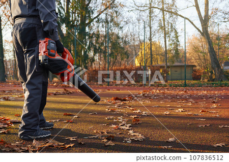 Unrecognizable male worker wearing blue uniform blowing out yellow leaves park area. Cropped view of strong man working with handheld leaf blower at outdoors tennis court. Seasonal work concept. Unrecognizable male worker wearing blue uniform blowing out yellow leaves park area. Cropped view of strong man working with handheld leaf blower at outdoors tennis court. Seasonal work concept. 107836512