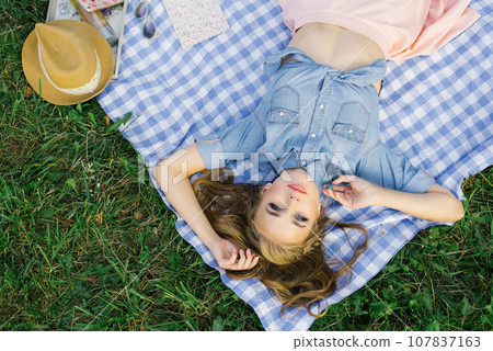 Stylish young woman with bright makeup in a denim shirt is lying on her back on a plaid in the open air and resting 107837163