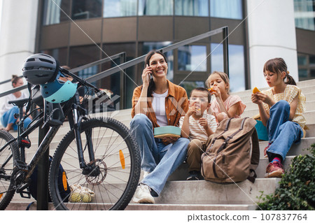 Children and mother have healthy snack in city after school. The family sitting on concrete steps in city, resting and eating home made snack, while mom is on the phone. Children and mother have healthy snack in city after school. The family sitting on concrete steps in city, resting and eating home made snack, while mom is on the phone. 107837764