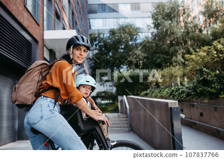 Mother carring her son on a child bike carrier, seat, both wearing helmets. Mom commuting with a young child through the city on a bicycle. 107837766
