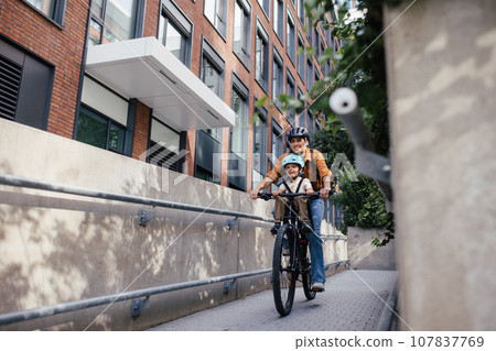 Mother carring her son on a child bike carrier, seat, both wearing helmets. Mom commuting with a young child through the city on a bicycle. 107837769