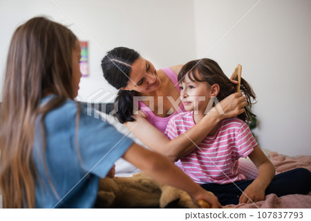 Mother with two daughters on a bed in the bedroom. Mother combs daughter's long hair with comb.Concept of Mother's Day and maternal love. 107837793