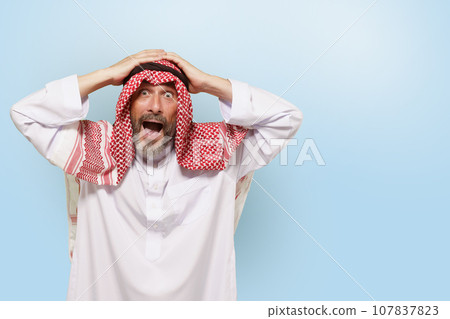 Excited Middle Eastern Arabian man, or Muslim, in traditional attire keffiyeh headscarf, with smile holds head. Joy and pride of Islamic people, cultural diversity and rich traditions of ethnic group Excited Middle Eastern Arabian man, or Muslim, in traditional attire keffiyeh headscarf, with smile holds head. Joy and pride of Islamic people, cultural diversity and rich traditions of ethnic group 107837823