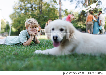 Children playing with a small puppy at a family garden party. Portrait of little boy lying on grass with Golden retriever puppy. 107837852
