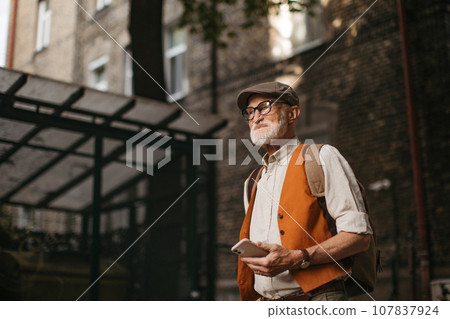Senior tourist exploring a new city and visiting interesting sites. Elderly man strolling through city, holding smartphone. Senior tourist exploring a new city and visiting interesting sites. Elderly man strolling through city, holding smartphone. 107837924