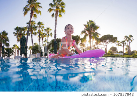 Girl floats on pink inflatable in the pool. Beach resort vacation by the sea. Winter and summer seaside resort holidays. Over-under underwater photography. 107837948