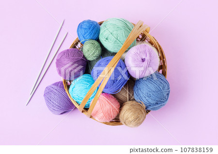 Lots of colorful knitting balls in a basket with knitting needles on a pastel purple background. Set for the hobby of knitting warm clothes or toys. Concept needlework. Flat lay Lots of colorful knitting balls in a basket with knitting needles on a pastel purple background. Set for the hobby of knitting warm clothes or toys. Concept needlework. Flat lay 107838159